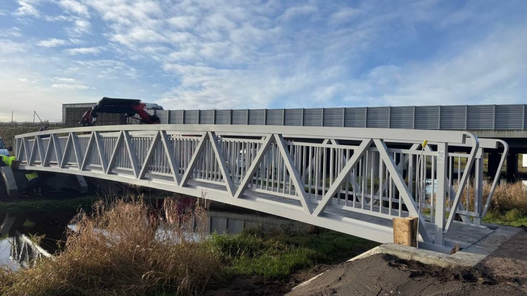 Light grey aluminium truss footbridge over a ditch beside a road with noise barriers