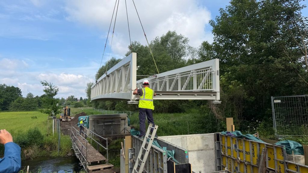 Worker guiding an aluminium footbridge span as a crane lowers it onto the abutments