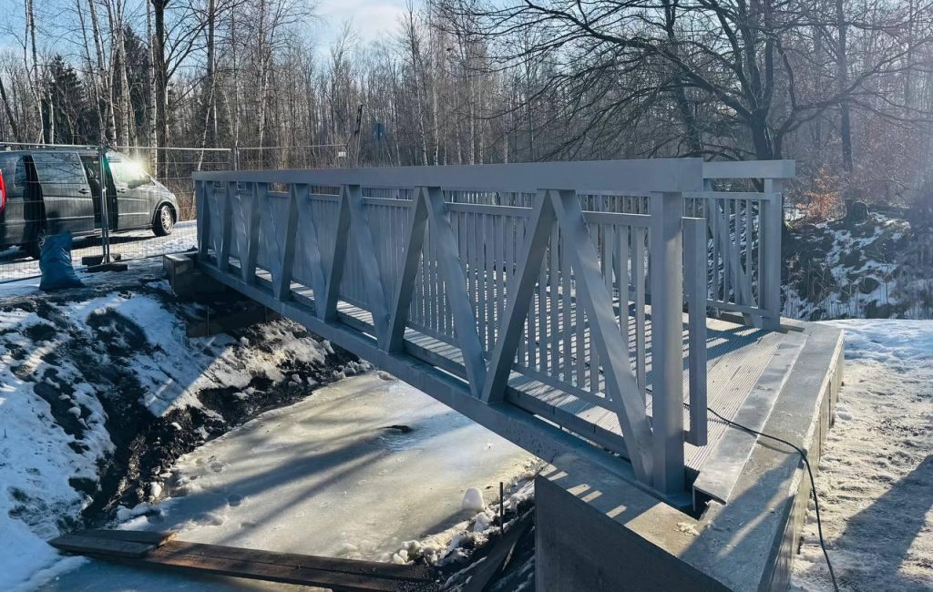 Grey aluminium truss footbridge over a frozen stream in a snow-covered birch forest