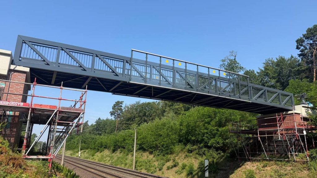 Dark grey aluminium footbridge over an electrified railway with scaffolded brick abutments