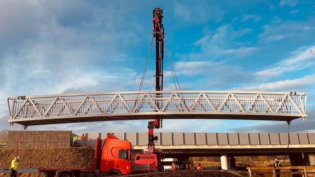 Aluminium truss bridge span suspended by a crane above a road during installation