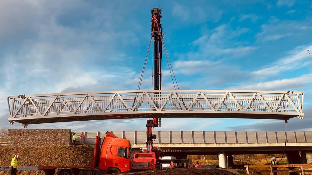 Aluminium truss bridge span suspended by a crane above a road during installation