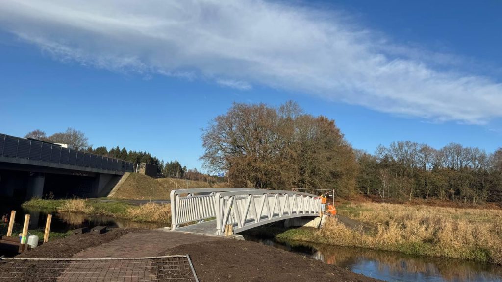 Light aluminium arched footbridge over a stream next to a motorway viaduct