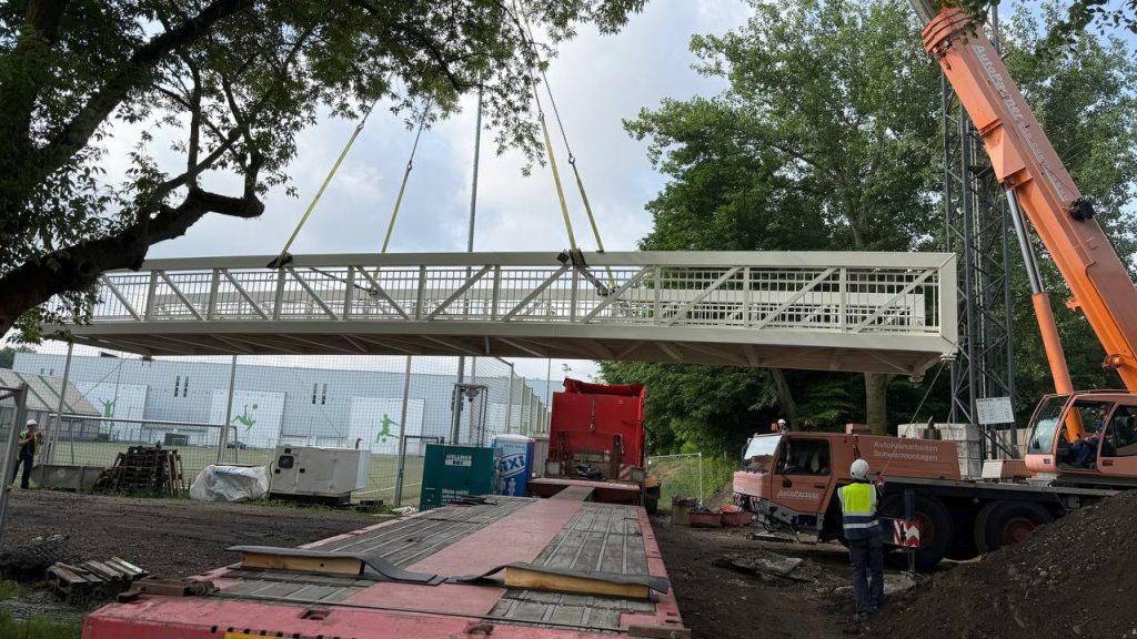 Cream aluminium footbridge span lifted by a mobile crane over a sports ground