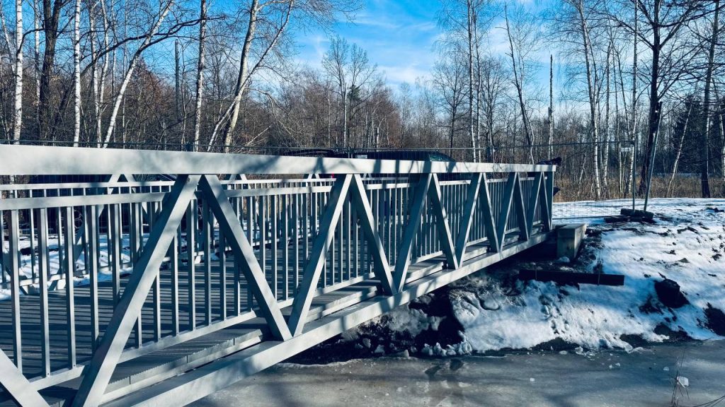 Light grey aluminium truss footbridge over frozen water in a birch forest